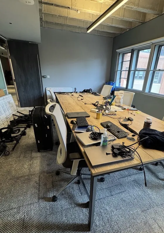 The old office before the move: a long table crowded with laptops, cables and keyboards, with a suitcase, whiteboards and equipment on the floor
